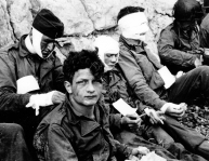 Wounded members of the 3rd Battalion, 16th Infantry Regiment receive cigarettes and food in a sheltered stretch of beach at the 6th Naval Beach Battalion’s medical aid station at the foot of chalk cliffs in the “Fox Red” sector of “Omaha” beach on the morning of D-Day. The soldier looking at the camera is identified as Private Nicholas A. Fina, 22-years old, of 'I' Company and New York state. (NARA 111-SC-189910)